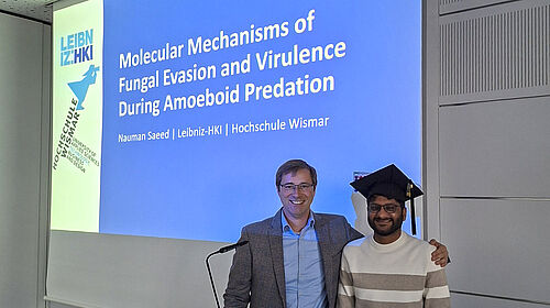 Doktor-Nauman Der Professor steht neben dem Doktoranden und hat seinen linken Arm auf dessen Schulter gelegt. Nauman Saeed hat einen Doiktorhut auf. Hinter den beiden ist auf einer Präsentationsfolie der Titel der Arbeit zu sehen.