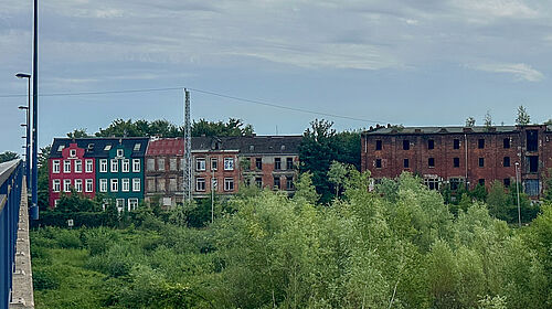 barrierefrei: Es ist ein verfallenes mehrstöckiges Backsteingebäude mit teilweise zerstörten Fenstern und angrenzenden bunten Wohnhäusern zu sehen - vor bewölktem Himmel und grüner Vegetation im Vordergrund.