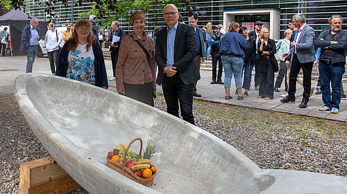 Es ist ein großer Löffel zu sehen, der auf einem Kieselboden liegt. Dahinter stehen die drei PErsonen. Noch weiter hinten stehen zahlrieche Gäste.
