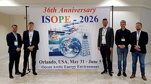 Die Mitglieder des Forschungsteams stehen links und rechts neben einem Ankündigungsbanner der ISOPE-Konferenz.