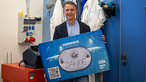 Der Mann steht in einem Labor und hält eine rund ein Meter breite Karte in der Hand. Sie sieht wie eine Dauerkarte aus, auf der in der Mitte "Danke" steht.