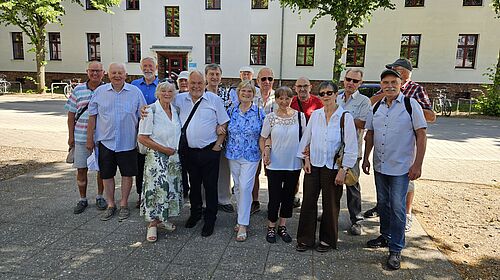 Eine Personengruppe steht bei sonnigem Wetter vor einem Gebäude.