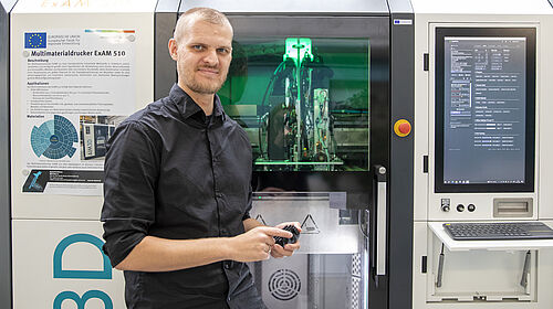 Ein Mann in schwarzem Hemd steht vor einem Multimaterialdrucker, einem großem Gerät, einem Schrank ähnlich, mit beleuchtetem Innenraum und großem Bildschirm rechts.